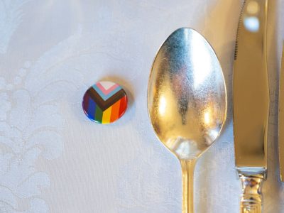 A Progress Pride pin badge beside silver cutlery on a white tablecloth, promotional image for the Inner Temple and LGBTQ+ Society Dinner