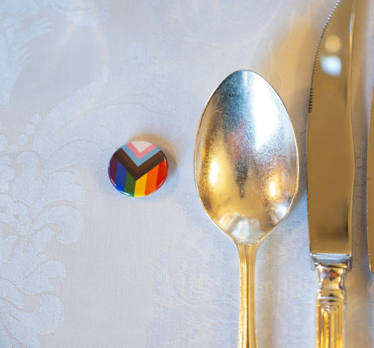 A Progress Pride pin badge beside silver cutlery on a white tablecloth, promotional image for the Inner Temple and LGBTQ+ Society Dinner